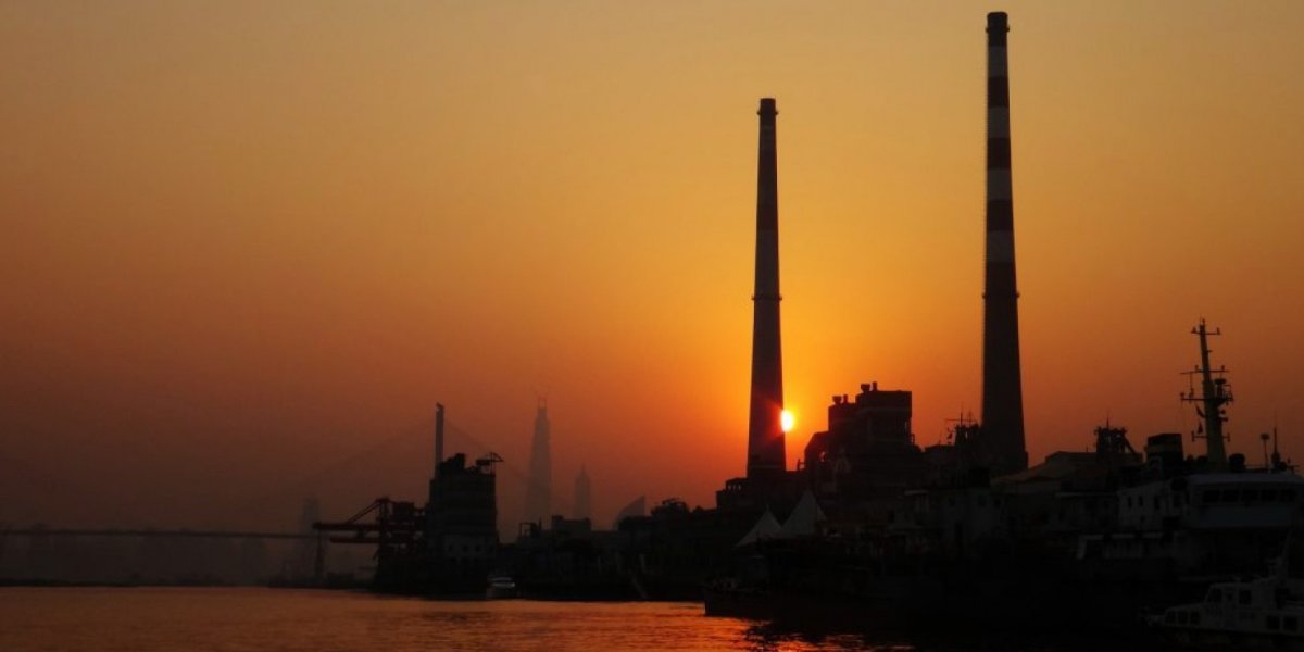 Bildet viser to høye fabrikkpiper i solnegang ved en sjø.
